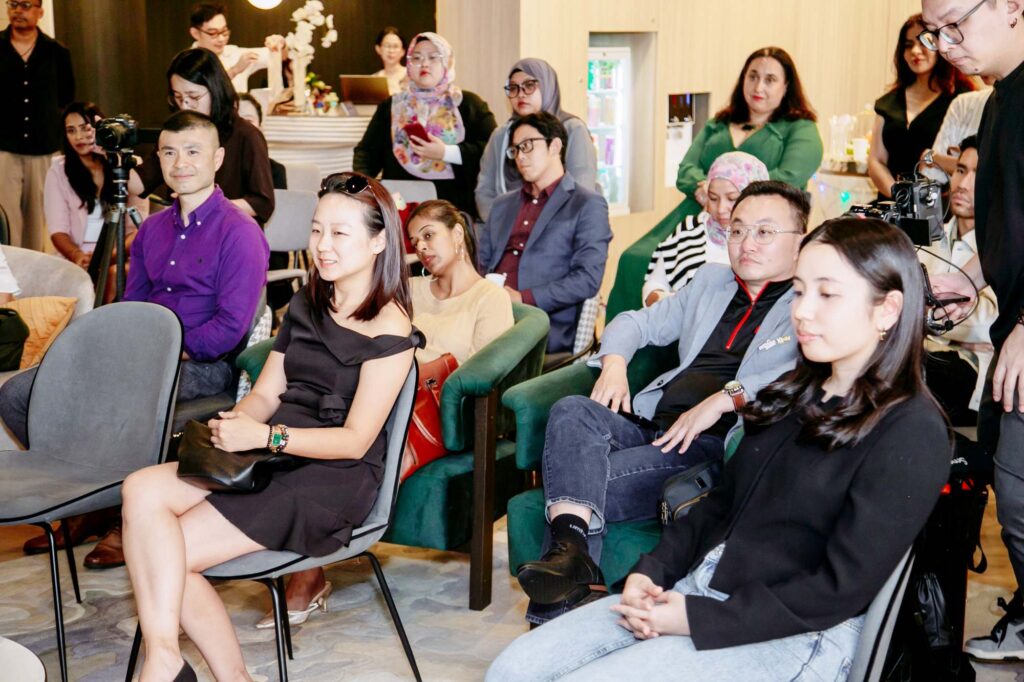 Audience gathered at an indoor event: seated attendees in front and people standing with cameras in the background, busy with photos.